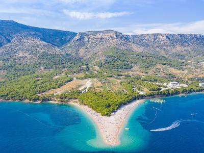 An aerial view of the distinctive golden, horn-shaped beach (Zlatni rat) jutting into the blue Adriatic Sea, with a large, forested mountain ridge rising directly behind it.