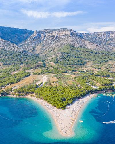 An aerial view of the distinctive golden, horn-shaped beach (Zlatni rat) jutting into the blue Adriatic Sea, with a large, forested mountain ridge rising directly behind it.