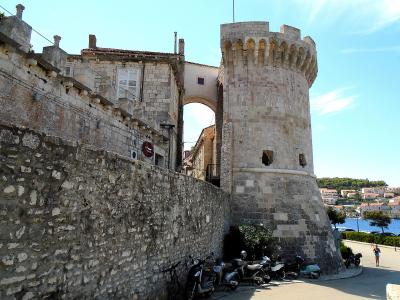 A stone medieval tower with crenellations and an arched gateway connecting to a fortified wall, with parked scooters and a coastal town view in the background.