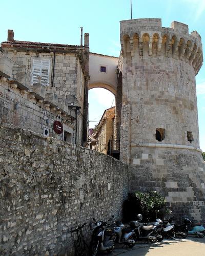 A stone medieval tower with crenellations and an arched gateway connecting to a fortified wall, with parked scooters and a coastal town view in the background.