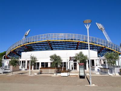 The image shows a modern outdoor tennis stadium with blue seating, a curved roof structure, and floodlights, featuring a white building base with signage for "24" and a Coca-Cola vending machine.
