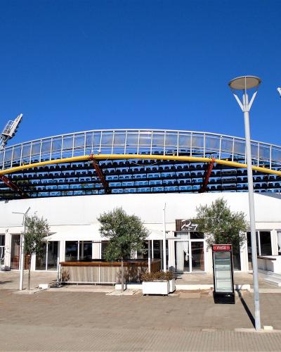 The image shows a modern outdoor tennis stadium with blue seating, a curved roof structure, and floodlights, featuring a white building base with signage for "24" and a Coca-Cola vending machine.