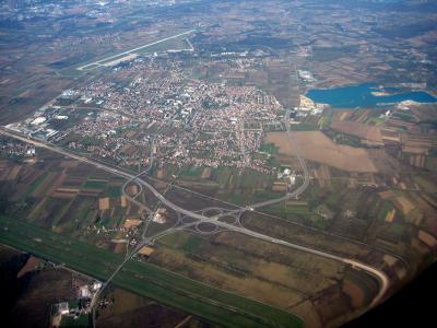 An aerial photograph showing the town of Velika Gorica, featuring a large highway interchange in the foreground, a dense urban area with red-roofed buildings, an airport runway in the upper left, and a lake to the right.