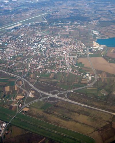 An aerial photograph showing the town of Velika Gorica, featuring a large highway interchange in the foreground, a dense urban area with red-roofed buildings, an airport runway in the upper left, and a lake to the right.