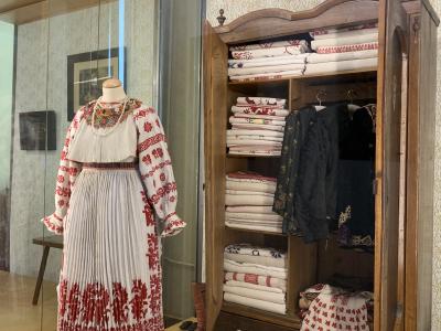 A traditional Croatian folk costume displayed on a mannequin next to an open wooden wardrobe filled with folded textiles and hanging garments inside a museum exhibit.