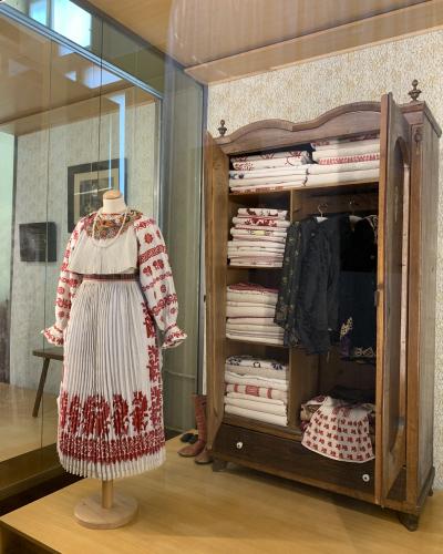 A traditional Croatian folk costume displayed on a mannequin next to an open wooden wardrobe filled with folded textiles and hanging garments inside a museum exhibit.