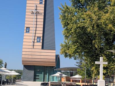 A modern church building with a tall, rectangular bell tower featuring horizontal beige and dark grey cladding, three visible bells, and a large metal cross mounted on the facade, situated next to a paved plaza with outdoor seating and a stone cross 