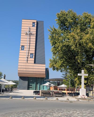 A modern church building with a tall, rectangular bell tower featuring horizontal beige and dark grey cladding, three visible bells, and a large metal cross mounted on the facade, situated next to a paved plaza with outdoor seating and a stone cross 