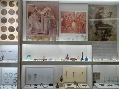 The image shows a museum display case containing ancient artifacts including coins, glass vessels, jewelry, and framed wall paintings or frescoes, with a label reading "METIKA COSMETICS" visible on the right side.