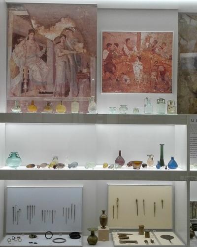 The image shows a museum display case containing ancient artifacts including coins, glass vessels, jewelry, and framed wall paintings or frescoes, with a label reading "METIKA COSMETICS" visible on the right side.
