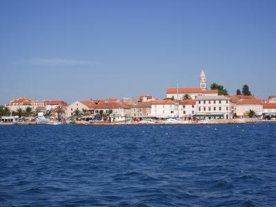 A coastal town scene with white buildings, terracotta roofs, a prominent church tower in the background, boats in the harbor, and blue sea in the foreground under a clear sky.