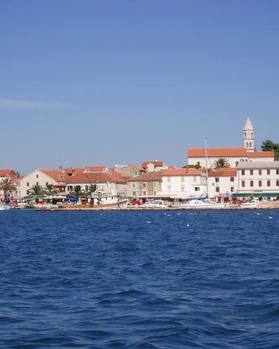 A coastal town scene with white buildings, terracotta roofs, a prominent church tower in the background, boats in the harbor, and blue sea in the foreground under a clear sky.