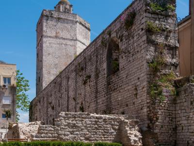 A view of ancient stone ruins and a tall tower with a dome against a blue sky, featuring layered stone walls and vegetation growing from the masonry.