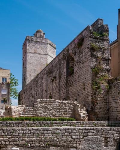A view of ancient stone ruins and a tall tower with a dome against a blue sky, featuring layered stone walls and vegetation growing from the masonry.