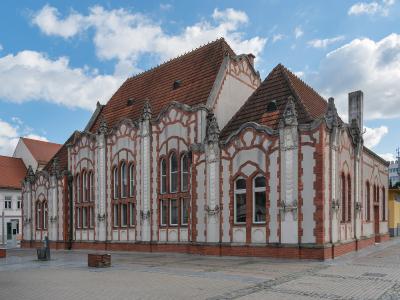 A red and white brick building with ornate Art Nouveau architectural details, including decorative gables and arched windows, situated on a paved public square under a blue sky.