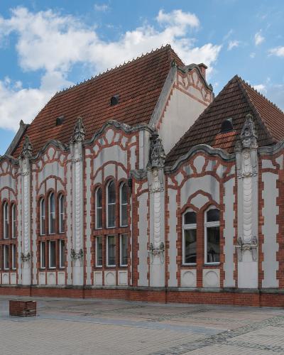 A red and white brick building with ornate Art Nouveau architectural details, including decorative gables and arched windows, situated on a paved public square under a blue sky.