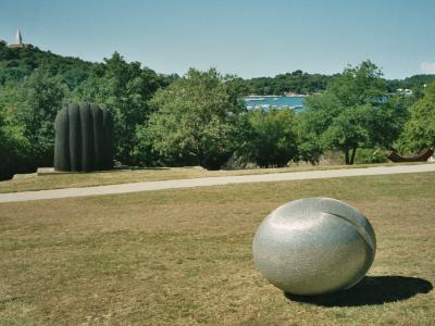 An outdoor park scene featuring large abstract sculptures, including a prominent silver sphere in the foreground and a dark ribbed column in the mid-ground, with a coastal view and a church steeple on a hill in the background.