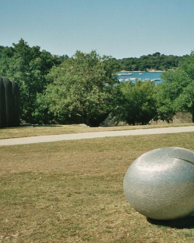 An outdoor park scene featuring large abstract sculptures, including a prominent silver sphere in the foreground and a dark ribbed column in the mid-ground, with a coastal view and a church steeple on a hill in the background.