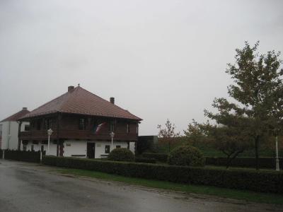 A two-story building with a white ground floor and a dark wooden upper story featuring a balcony, a red tiled roof, and a Croatian flag, situated next to a paved road and a hedge.