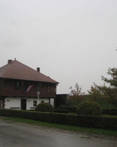 A two-story building with a white ground floor and a dark wooden upper story featuring a balcony, a red tiled roof, and a Croatian flag, situated next to a paved road and a hedge.