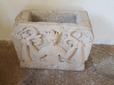 A stone architectural fragment featuring a relief carving of a crowned figure holding two serpents, resting on a stone floor against a plain wall.
