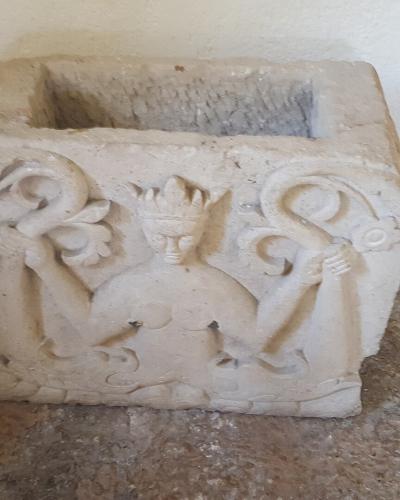 A stone architectural fragment featuring a relief carving of a crowned figure holding two serpents, resting on a stone floor against a plain wall.