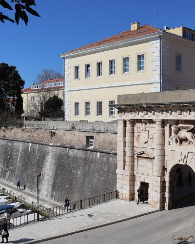 The image shows a large stone gatehouse with classical columns and a relief of a winged lion, situated next to a harbor filled with boats and a high stone seawall.