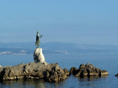 A bronze statue of a standing female figure holding a bird, positioned on a large rock formation extending into the sea with a coastal town and mountains in the background.
