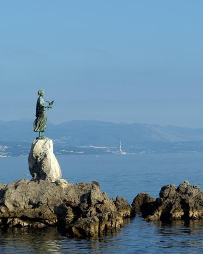 A bronze statue of a standing female figure holding a bird, positioned on a large rock formation extending into the sea with a coastal town and mountains in the background.