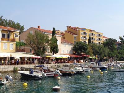 A view of a Croatian coastal harbor with small boats moored in the foreground, a stone promenade lined with outdoor cafes and restaurants, and multi-story buildings in the background, including a yellow hotel with "Hotel Selce" signage on its roof