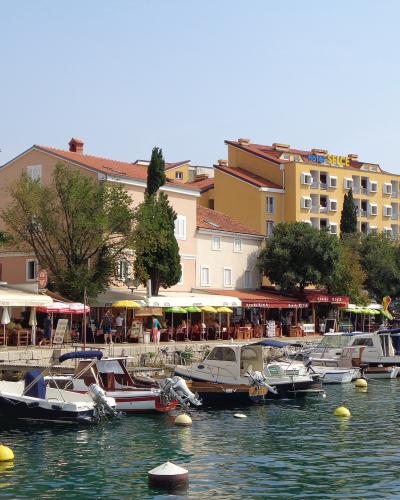 A view of a Croatian coastal harbor with small boats moored in the foreground, a stone promenade lined with outdoor cafes and restaurants, and multi-story buildings in the background, including a yellow hotel with "Hotel Selce" signage on its roof