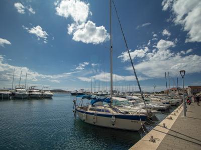 A sunny marina scene showing a paved waterfront promenade lined with docked sailboats and motor yachts under a blue sky with scattered clouds.