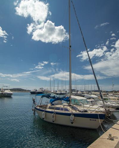 A sunny marina scene showing a paved waterfront promenade lined with docked sailboats and motor yachts under a blue sky with scattered clouds.