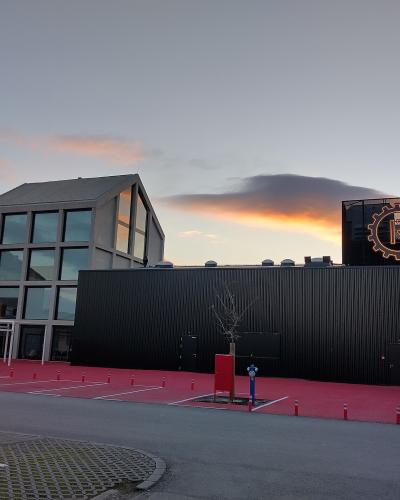 A modern building complex with a black corrugated metal facade and a distinctive illuminated gear-shaped logo containing the letter 'R' on the roof, set against a twilight sky.