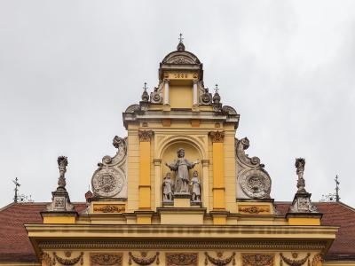 The image shows the ornate Baroque pediment of a yellow building with a central statue of a religious figure flanked by two smaller statues, set against a cloudy sky; the date "1888" is visible on the facade.