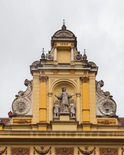 The image shows the ornate Baroque pediment of a yellow building with a central statue of a religious figure flanked by two smaller statues, set against a cloudy sky; the date "1888" is visible on the facade.