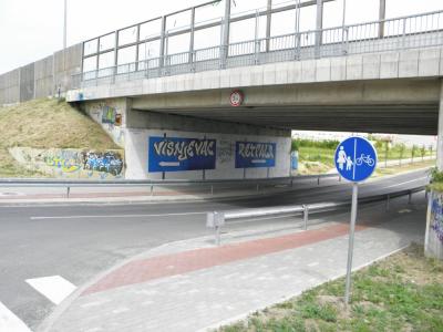 A concrete underpass beneath a bridge with blue directional signs reading "VIŠNJEGAC" and "RETFAŁA" pointing in opposite directions, alongside a blue pedestrian and bicycle path sign.
