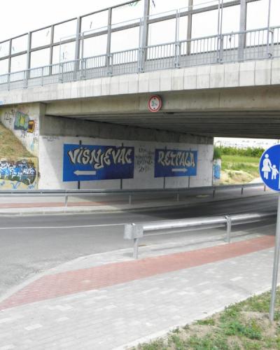 A concrete underpass beneath a bridge with blue directional signs reading "VIŠNJEGAC" and "RETFAŁA" pointing in opposite directions, alongside a blue pedestrian and bicycle path sign.