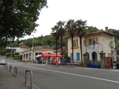 A street scene showing a road with a "PARKING" sign, a small kiosk with red Coca-Cola umbrellas, and a yellow building with palm trees.