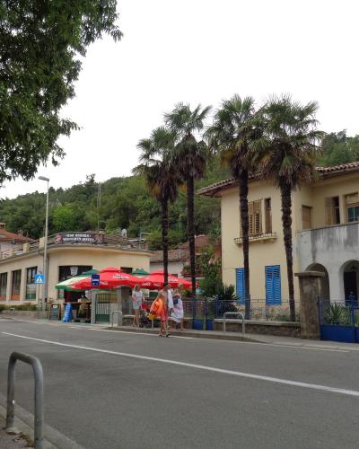A street scene showing a road with a "PARKING" sign, a small kiosk with red Coca-Cola umbrellas, and a yellow building with palm trees.