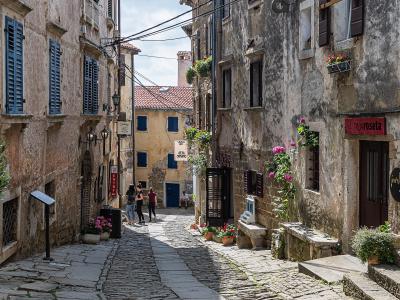 A narrow, cobblestone street in a historic European town, featuring old stone buildings with shuttered windows, potted flowers, and several signs including "GALERIJA roseta" and "SETA STUDIO".