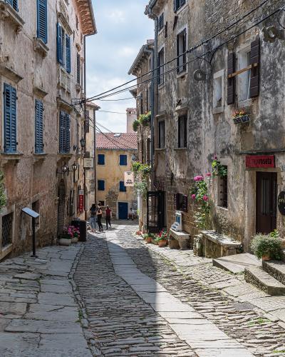 A narrow, cobblestone street in a historic European town, featuring old stone buildings with shuttered windows, potted flowers, and several signs including "GALERIJA roseta" and "SETA STUDIO".