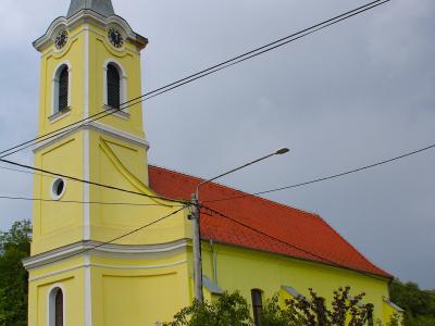 A yellow church with a tall steeple, red-tiled roof, and white trim, situated beside a road with trees and a wooden fence.
