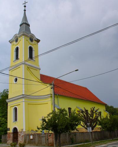 A yellow church with a tall steeple, red-tiled roof, and white trim, situated beside a road with trees and a wooden fence.