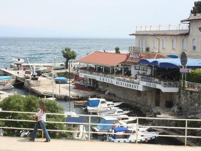 A waterfront restaurant with a red-tiled roof and blue awnings situated on a stone pier in a small harbor, with boats docked below and the sea in the background.
