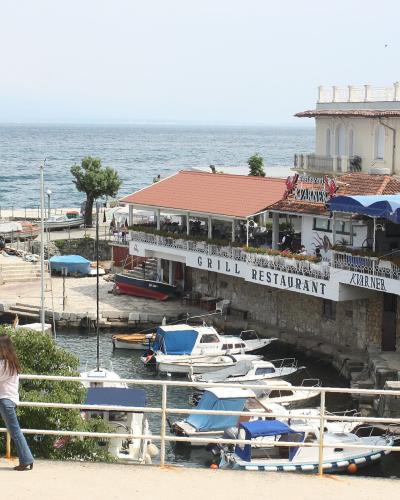 A waterfront restaurant with a red-tiled roof and blue awnings situated on a stone pier in a small harbor, with boats docked below and the sea in the background.