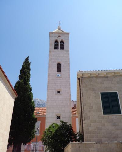 A tall, white stone bell tower with a cross on top, arched belfry openings, and decorative cross-shaped vents, set against a blue sky with surrounding buildings and a cypress tree.