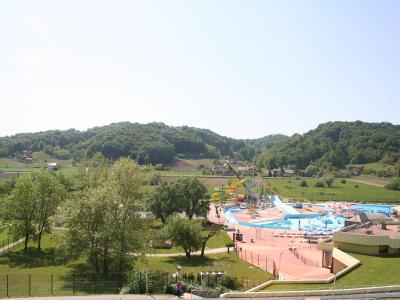 A wide-angle view of a large outdoor thermal water park featuring multiple colorful water slides, swimming pools, and surrounding green hills with scattered houses.