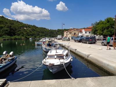 A sunny waterfront scene showing a paved promenade along a calm harbor with several small boats moored, cars parked along the edge, and a green hillside in the background under a blue sky with clouds.