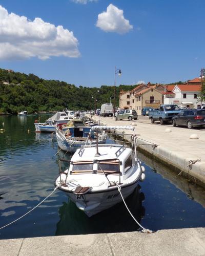 A sunny waterfront scene showing a paved promenade along a calm harbor with several small boats moored, cars parked along the edge, and a green hillside in the background under a blue sky with clouds.